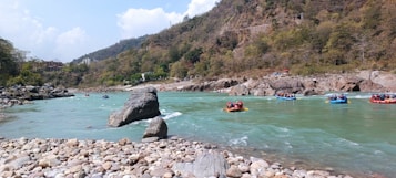 A scenic river landscape with groups of people rafting on turquoise water. The river is surrounded by rocky terrain and lush green hills in the background, with a cloudy sky above. Large rocks are scattered along the riverbanks.