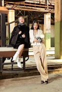 Two women in fashionable attire pose in an industrial setting. One wears a black outfit with a polka-dot headscarf and white boots, sitting on a metal structure. The other stands nearby in a cream suit, holding a vintage camera. The background features rustic metal beams and concrete columns, creating a contrast with their stylish looks.