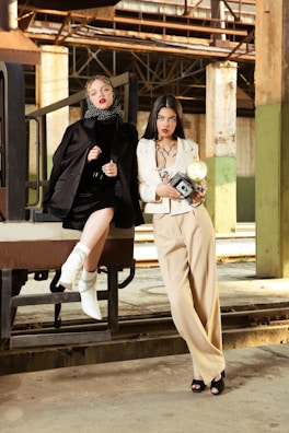 Two women in fashionable attire pose in an industrial setting. One wears a black outfit with a polka-dot headscarf and white boots, sitting on a metal structure. The other stands nearby in a cream suit, holding a vintage camera. The background features rustic metal beams and concrete columns, creating a contrast with their stylish looks.