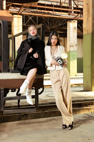 Two women in fashionable attire pose in an industrial setting. One wears a black outfit with a polka-dot headscarf and white boots, sitting on a metal structure. The other stands nearby in a cream suit, holding a vintage camera. The background features rustic metal beams and concrete columns, creating a contrast with their stylish looks.