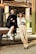 Two women in fashionable attire pose in an industrial setting. One wears a black outfit with a polka-dot headscarf and white boots, sitting on a metal structure. The other stands nearby in a cream suit, holding a vintage camera. The background features rustic metal beams and concrete columns, creating a contrast with their stylish looks.