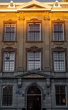 A grand building with classical architectural elements, featuring large, symmetrical windows and detailed stonework. The facade includes Roman numerals 'ANNO MCMX' and the name 'Dutch Golden Hotel' above the entrance. A warm, golden light illuminates the upper portion.