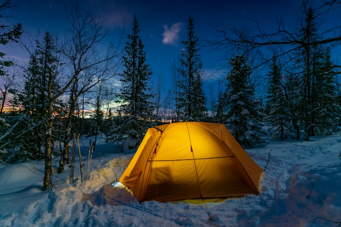 Orange dome tent glowing from a light inside, pitched on deep snow among spruce and birch trees under a blue evening sky