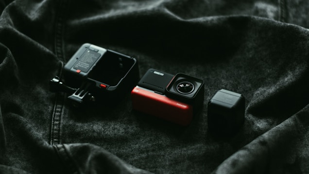 A close-up shot of a modular action camera disassembled into three main parts, placed on a dark, textured fabric. The components include the main body with a red and black finish, a lens module, and a frame case.