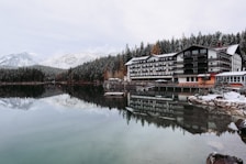 A tranquil lakeside scene features a large, multi-story hotel with a wooden exterior. The building is reflected in the calm, clear water of the lake. Snow-dusted pine forests and snow-capped mountains form a picturesque backdrop. The leafless trees and snow-covered ground suggest a cold, winter setting.