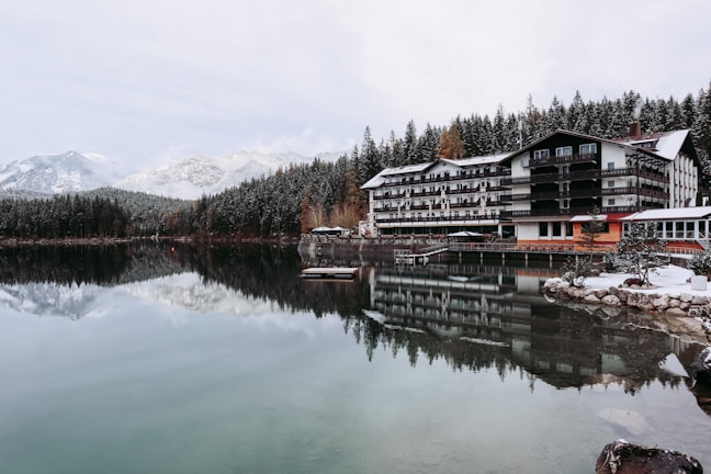 A tranquil lakeside scene features a large, multi-story hotel with a wooden exterior. The building is reflected in the calm, clear water of the lake. Snow-dusted pine forests and snow-capped mountains form a picturesque backdrop. The leafless trees and snow-covered ground suggest a cold, winter setting.