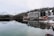 A tranquil lakeside scene features a large, multi-story hotel with a wooden exterior. The building is reflected in the calm, clear water of the lake. Snow-dusted pine forests and snow-capped mountains form a picturesque backdrop. The leafless trees and snow-covered ground suggest a cold, winter setting.