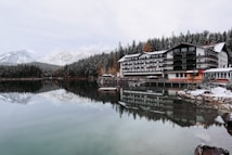A tranquil lakeside scene features a large, multi-story hotel with a wooden exterior. The building is reflected in the calm, clear water of the lake. Snow-dusted pine forests and snow-capped mountains form a picturesque backdrop. The leafless trees and snow-covered ground suggest a cold, winter setting.