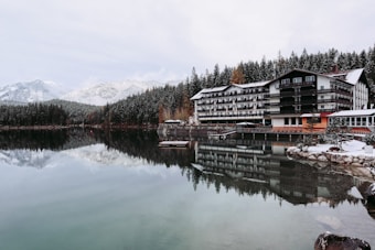 A tranquil lakeside scene features a large, multi-story hotel with a wooden exterior. The building is reflected in the calm, clear water of the lake. Snow-dusted pine forests and snow-capped mountains form a picturesque backdrop. The leafless trees and snow-covered ground suggest a cold, winter setting.