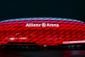 A large stadium facade illuminated in vibrant red lighting. The structure has a modern, geometric pattern with the name 'Allianz Arena' prominently displayed in white letters. The surrounding area is dark, highlighting the striking visual effect of the stadium's illumination.