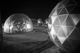 Several geodesic dome structures with illuminated interiors are arranged on a barren landscape at night. The domes are constructed with a pattern of triangular panels, reflecting light and casting shadows. The scene suggests a futuristic or extraterrestrial environment.
