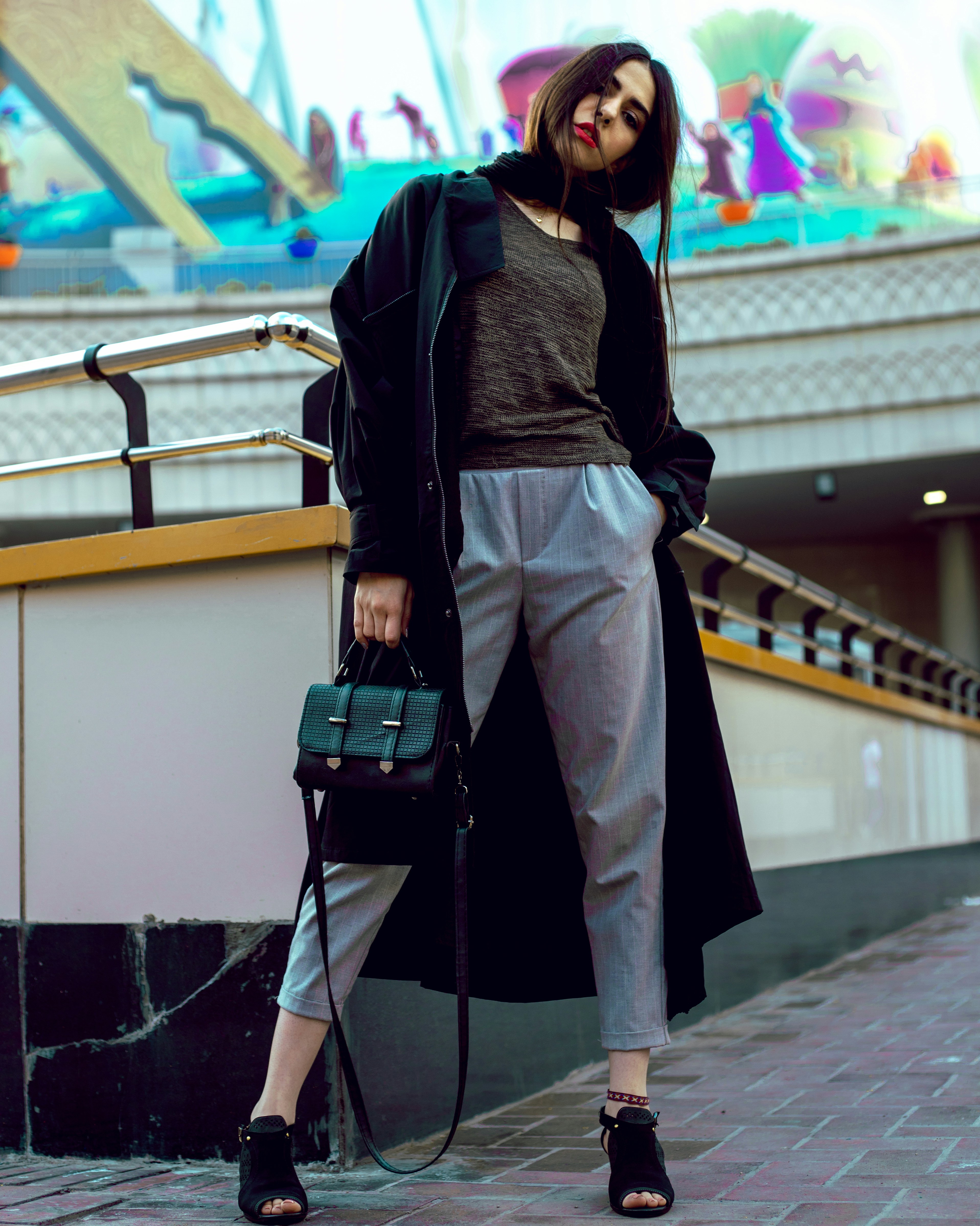 A fashionably dressed woman stands confidently on a city sidewalk, wearing a long dark coat, a casual top, and light grey pants. She holds a stylish handbag and wears open-toe black sandals. The background features a whimsical mural with colorful abstract shapes and playful figures, giving a lively contrast to her urban attire.