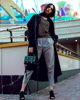 A fashionably dressed woman stands confidently on a city sidewalk, wearing a long dark coat, a casual top, and light grey pants. She holds a stylish handbag and wears open-toe black sandals. The background features a whimsical mural with colorful abstract shapes and playful figures, giving a lively contrast to her urban attire.
