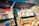 A vibrant outdoor bookstall displays an array of books, magazines, and artwork. The books and magazines are arranged neatly on open display under large open lids, with some items hanging on strings. The scene suggests a sunny day with shadows cast by nearby trees and structures.