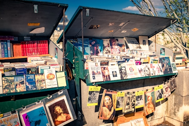 A vibrant outdoor bookstall displays an array of books, magazines, and artwork. The books and magazines are arranged neatly on open display under large open lids, with some items hanging on strings. The scene suggests a sunny day with shadows cast by nearby trees and structures.