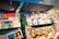A vibrant outdoor bookstall displays an array of books, magazines, and artwork. The books and magazines are arranged neatly on open display under large open lids, with some items hanging on strings. The scene suggests a sunny day with shadows cast by nearby trees and structures.