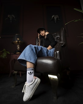 A person wearing a black top, wide-legged jeans, a cap, and white sneakers with branded socks lounges casually in a dark, sophisticated room. They sit in a luxurious leather chair, beside a small side table with a designer lamp. Two framed ram skulls adorn the walls, adding a rustic touch to the decor.