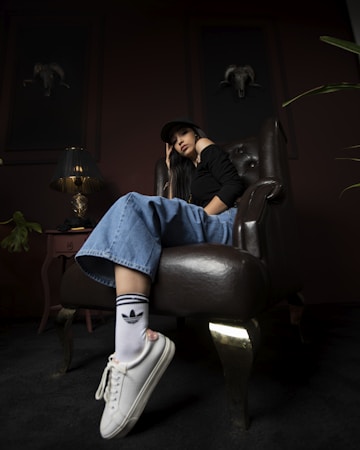 A person wearing a black top, wide-legged jeans, a cap, and white sneakers with branded socks lounges casually in a dark, sophisticated room. They sit in a luxurious leather chair, beside a small side table with a designer lamp. Two framed ram skulls adorn the walls, adding a rustic touch to the decor.