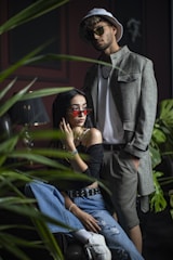 A man and a woman are fashionably dressed in a trendy and modern style, surrounded by lush green plants. The woman is sitting on a chair, wearing off-the-shoulder black top, blue jeans, and white sneakers, accessorized with a belt and sunglasses. The man stands behind her, wearing a light gray jacket, shorts, a white t-shirt, sunglasses, and a hat. The setting is softly lit, creating a stylish, urban atmosphere.