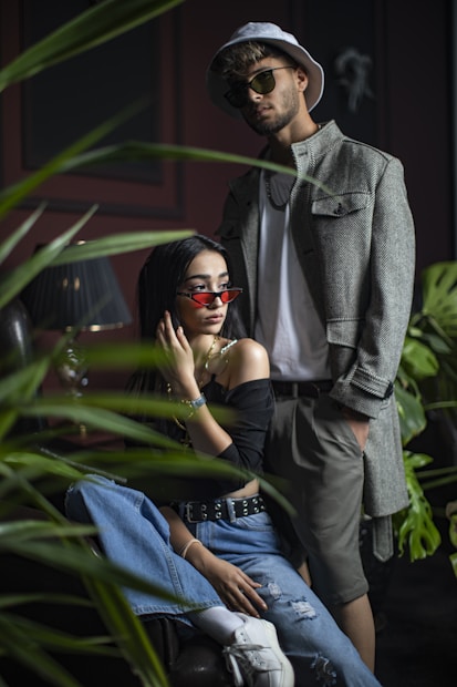 A man and a woman are fashionably dressed in a trendy and modern style, surrounded by lush green plants. The woman is sitting on a chair, wearing off-the-shoulder black top, blue jeans, and white sneakers, accessorized with a belt and sunglasses. The man stands behind her, wearing a light gray jacket, shorts, a white t-shirt, sunglasses, and a hat. The setting is softly lit, creating a stylish, urban atmosphere.