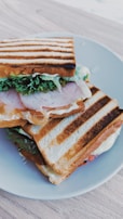 A toasted sandwich placed on a white plate, featuring layers of neatly stacked grilled bread filled with lettuce, slices of meat, and melted cheese. The grill marks are distinct across the top slice of bread, and the contents spill slightly over the sides.