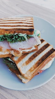 A toasted sandwich placed on a white plate, featuring layers of neatly stacked grilled bread filled with lettuce, slices of meat, and melted cheese. The grill marks are distinct across the top slice of bread, and the contents spill slightly over the sides.