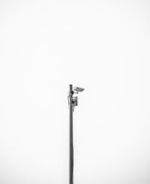 A minimalistic image featuring a single security camera mounted on a tall metal pole against a plain white background. The setup is centralized and the black and white color scheme adds a stark contrast, emphasizing the industrial design of the camera.