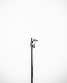 A minimalistic image featuring a single security camera mounted on a tall metal pole against a plain white background. The setup is centralized and the black and white color scheme adds a stark contrast, emphasizing the industrial design of the camera.