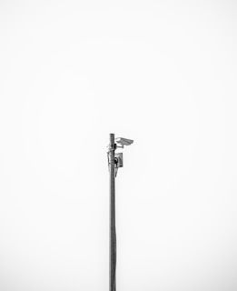 A minimalistic image featuring a single security camera mounted on a tall metal pole against a plain white background. The setup is centralized and the black and white color scheme adds a stark contrast, emphasizing the industrial design of the camera.