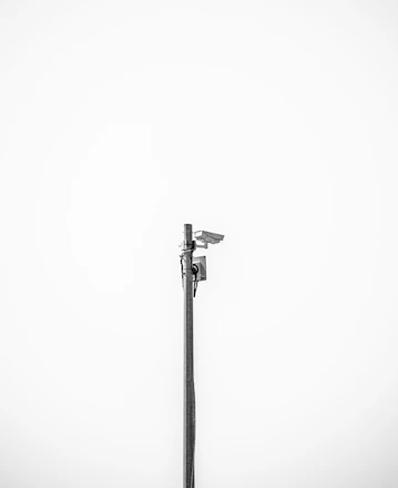 A minimalistic image featuring a single security camera mounted on a tall metal pole against a plain white background. The setup is centralized and the black and white color scheme adds a stark contrast, emphasizing the industrial design of the camera.