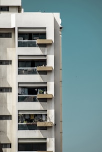 A modern high-rise apartment building with multiple balconies, some featuring glass railings. Various personal items and laundry can be seen on the balconies. The building exterior is white with some weathering visible. The sky in the background is clear and light blue.