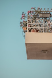 A balcony set against a clear blue sky, featuring a variety of potted plants and flowers. The railing has a lattice design with some green foliage and vibrant pink flowers spilling over it. The structure beneath the balcony is an off-white color with a lamp attached.