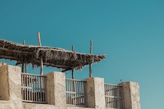A rustic structure features wooden poles and a roof made from dried palm leaves, set against a clear blue sky. The building has stone columns and wooden railings, suggesting a traditional or coastal architecture style.