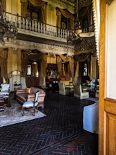 A lavishly decorated interior with high ceilings and ornate molding. Chandeliers hang from the ceiling, and the room includes antique-style furniture such as sofas and armchairs with elegant fabric. Heavy drapes and gold accents add to the opulent atmosphere. The floor features dark wood with a herringbone pattern, and various decorative items are placed throughout.