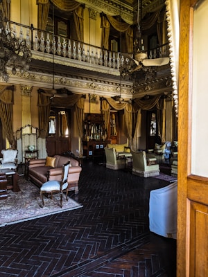 A lavishly decorated interior with high ceilings and ornate molding. Chandeliers hang from the ceiling, and the room includes antique-style furniture such as sofas and armchairs with elegant fabric. Heavy drapes and gold accents add to the opulent atmosphere. The floor features dark wood with a herringbone pattern, and various decorative items are placed throughout.