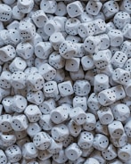 A large pile of white dice is scattered, each with black dots representing the numbers one through six. The dice are tightly packed together, creating a sense of randomness and abundance.