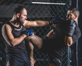 Two men are engaged in a martial arts sparring session inside a caged area. One man is delivering a knee strike towards the other, who is defending with gloves. Both are wearing sports attire and appear focused and intense.