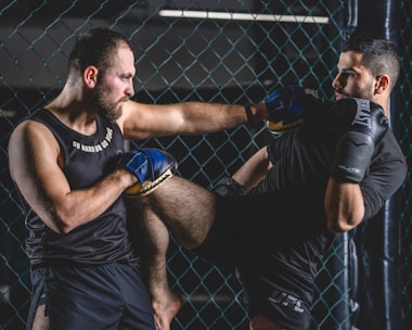 Two men are engaged in a martial arts sparring session inside a caged area. One man is delivering a knee strike towards the other, who is defending with gloves. Both are wearing sports attire and appear focused and intense.