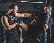 Two men are engaged in a martial arts sparring session inside a caged area. One man is delivering a knee strike towards the other, who is defending with gloves. Both are wearing sports attire and appear focused and intense.