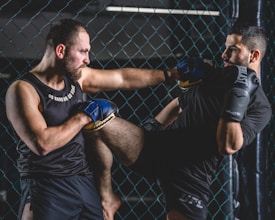 Two men are engaged in a martial arts sparring session inside a caged area. One man is delivering a knee strike towards the other, who is defending with gloves. Both are wearing sports attire and appear focused and intense.