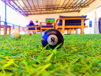 A close-up view of an 8-ball from a pool table lying on a green artificial turf. In the background, there are wooden chairs with navy blue cushions and two people sitting. The area appears to be a covered outdoor or semi-outdoor space with a roof and railings.