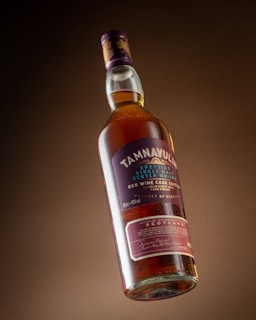 A bottle of Tamnavulin Speyside single malt Scotch whisky, featuring a red wine cask edition. The bottle is suspended at a slight angle with a rich amber liquid inside. The label on the bottle is prominently displayed, highlighting key details about the whisky, including its origin from Scotland and a purple and gold color scheme.