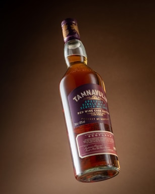 A bottle of Tamnavulin Speyside single malt Scotch whisky, featuring a red wine cask edition. The bottle is suspended at a slight angle with a rich amber liquid inside. The label on the bottle is prominently displayed, highlighting key details about the whisky, including its origin from Scotland and a purple and gold color scheme.