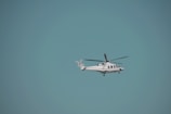 A helicopter is flying against a clear blue sky. The helicopter is white with red accents and has visible rotor blades. It is centered slightly to the right of the image with ample space surrounding it, highlighting the vastness of the sky.
