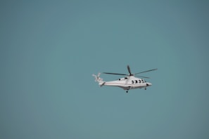 A helicopter is flying against a clear blue sky. The helicopter is white with red accents and has visible rotor blades. It is centered slightly to the right of the image with ample space surrounding it, highlighting the vastness of the sky.