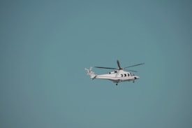 A helicopter is flying against a clear blue sky. The helicopter is white with red accents and has visible rotor blades. It is centered slightly to the right of the image with ample space surrounding it, highlighting the vastness of the sky.