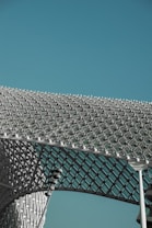 An intricate metal structure with a geometric, lattice-like design, forming a curved architectural feature against a clear blue sky. The structure appears to be made of numerous square and rectangular elements, interconnected to form a mesh.