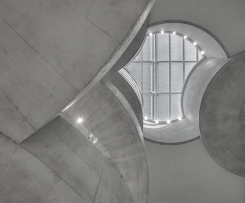 The interior of a modern architectural building is depicted with large, curved concrete structures that frame a circular skylight. The ceiling features a grid-like pattern, and small lights illuminate the curves, creating a futuristic atmosphere.