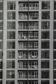 Tall apartment building with multiple floors and balconies filled with household items. Each floor has glass railings and sliding doors, with some balconies displaying clothes hanging to dry.