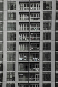 Tall apartment building with multiple floors and balconies filled with household items. Each floor has glass railings and sliding doors, with some balconies displaying clothes hanging to dry.